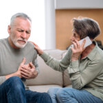 Elderly husband holding hand on chest, having heart attack, feeling unwell. Worried wife supporting partner call 911. Symptoms of myocardial infarction. Ambulance and immediate emergency help